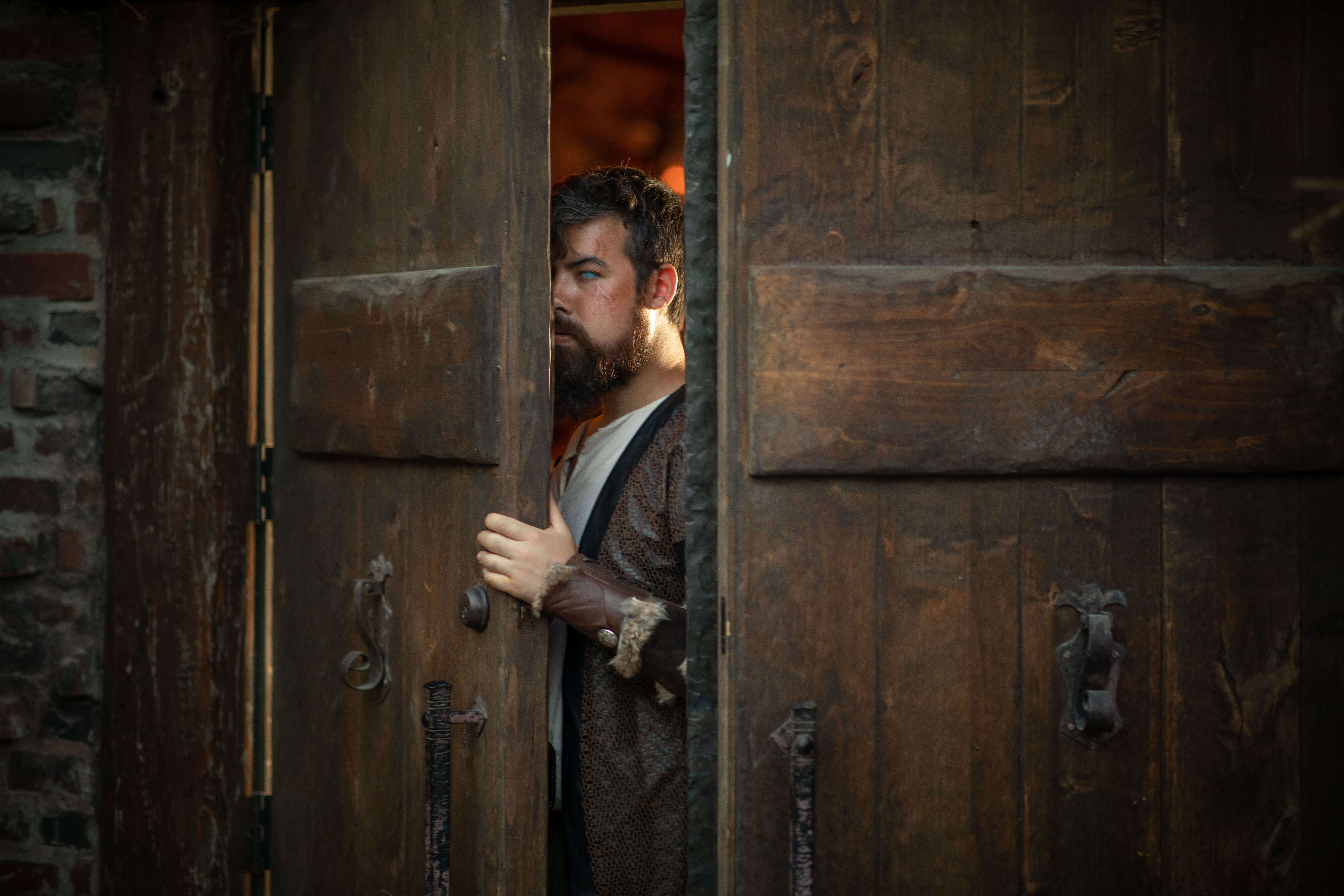 A human bartender in a Dungeons and Dragons tavern stands peering through a heavy set of double wooden doors